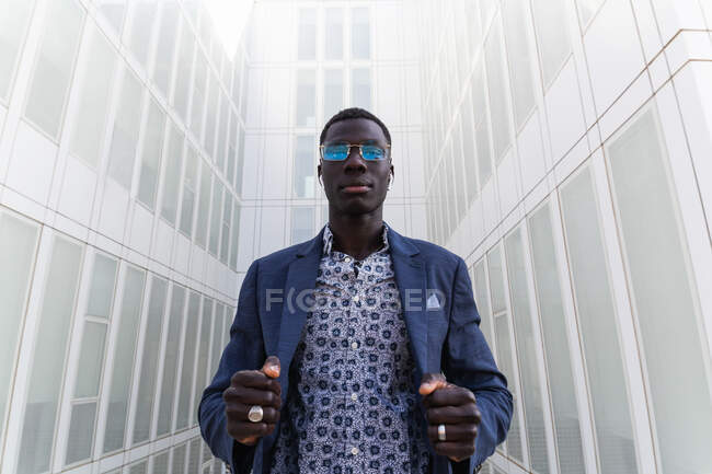 Low angle of stylish African American male entrepreneur listening to music in wireless earphones while standing in downtown and looking at camera — Stock Photo