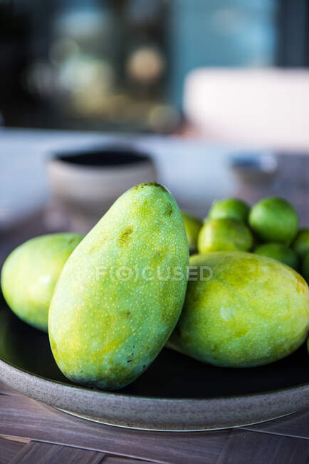 Exotic green mangoes on plate served on table with rip limes on sunny day in tropical resort - foto de stock