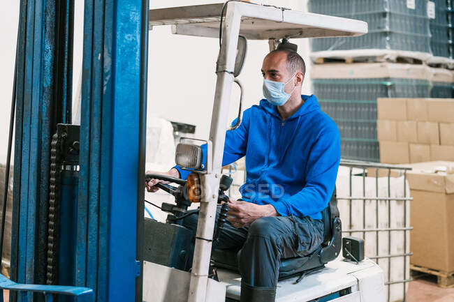 Male engineer in sterile mask driving vehicle and looking forward against cardboard boxes and bottles with beer in factory — Stock Photo