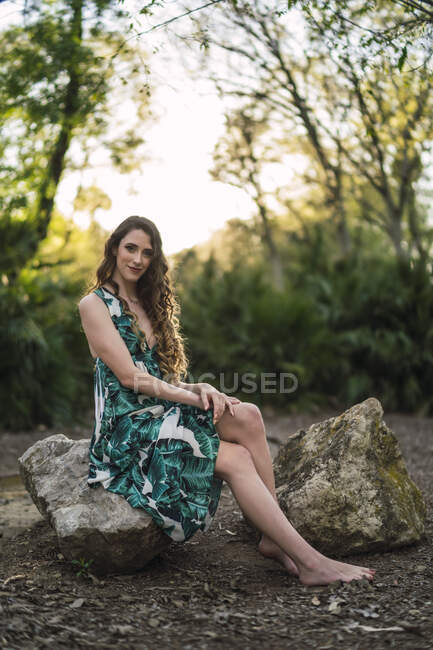 Contenuto del corpo pieno giovane femmina indossa maxi sundress alla moda e guardando la fotocamera con il sorriso mentre seduto su una grande pietra nel verdeggiante bosco soleggiato — Foto stock