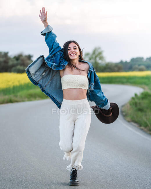 Comprimento total encantado jovem fêmea na roupa de verão elegante levantando braço e passeando na estrada de asfalto estreita que atravessa a área rural exuberante — Fotografia de Stock