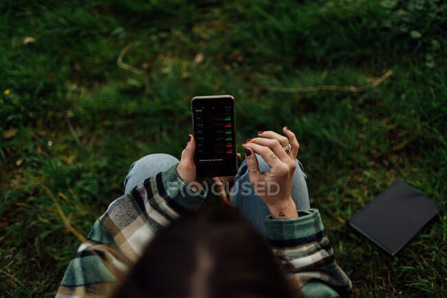 From above of crop anonymous female dealer touching screen of cellphone with graphs while sitting on meadow — Stock Photo
