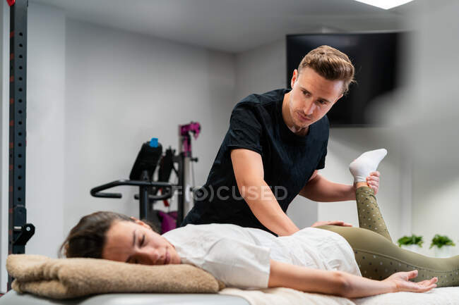 Fisioterapista maschio adulto che tocca la gamba della donna con gli occhi chiusi durante l'esame sul letto in ospedale — Foto stock