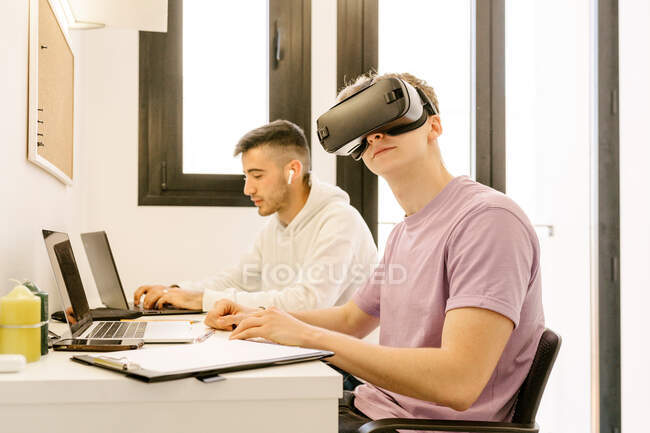 Side view of positive young male employee in casual clothes experiencing virtual reality in modern headset while working on laptop with smiling ethnic colleague — Stock Photo