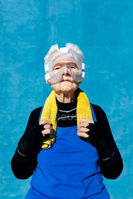 Serious mature female in activewear in protective boxing head guard with towel on shoulder standing on blue background and looking at camera — Stock Photo