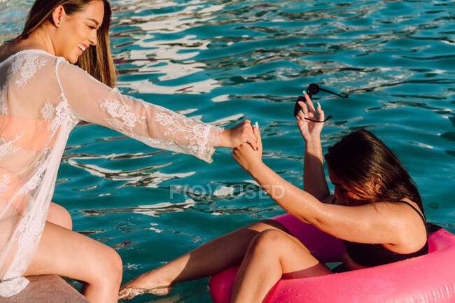 Vista lateral de sorrir viajante feminino no anel de borracha segurando a mão de amigo alegre na beira da piscina durante as férias — Fotografia de Stock