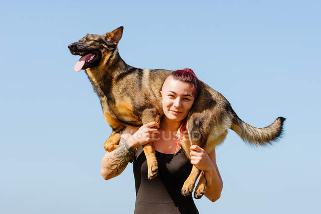 Atleta feminina tatuada com cão de raça pura bonito nos ombros olhando para a câmera no dia ensolarado — Fotografia de Stock