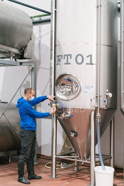 Vista lateral do trabalhador masculino em máscara estéril derramando cerveja fermentada de vaso de metal em vidro na fábrica — Fotografia de Stock