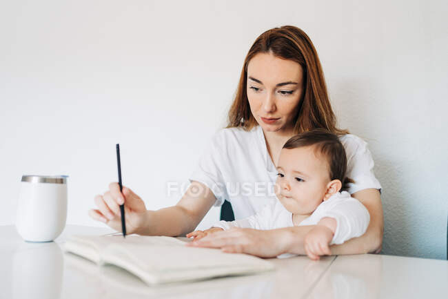 Positive young mother with adorable baby writing on diary while sitting together at desk in light room — Stock Photo