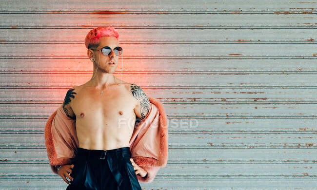 Jeune gay dans des lunettes de soleil modernes et veste de fourrure élégante avec des cheveux roses contre le mur gris — Photo de stock