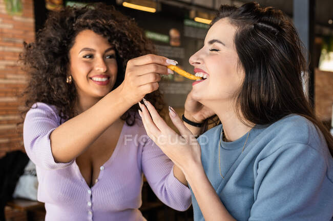 Joven hembra étnica positiva con pelo rizado oscuro alimentando hambrienta alegre amiga con apetitosas papas fritas en el restaurante - foto de stock