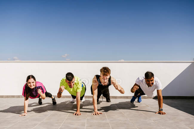 Grupo de cuerpo completo de alegres jóvenes amigos atléticos multirraciales en ropa deportiva haciendo ejercicio de tablón en la estera durante el entrenamiento en la terraza contra el cielo azul sin nubes - foto de stock