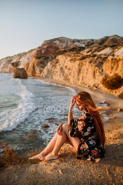 Женщина в мини-юбке сидит на грубом скалистом побережье и трогает длинные волосы под ясным голубым небом в Fyriplaka Милош — стоковое фото