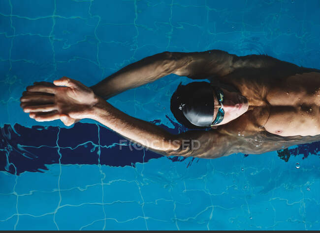 Вид сверху спортсмена-мужчины в купальной шапке с поднятыми руками, плавающего на спине в бассейне во время тренировки — стоковое фото