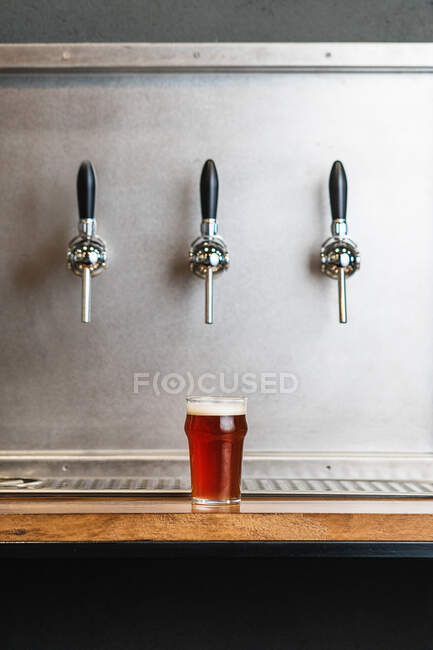 Cerveja com espuma em jarro de vidro contra linha de torneiras em bar no fundo cinza — Fotografia de Stock