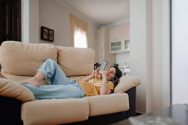 Vista lateral de la joven hembra tomando autorretrato en el teléfono celular mientras se acuesta en el sofá en la sala de estar - foto de stock