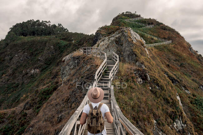 Back view of unrecognizable male tourist with backpack and hat standing on wooden pathway in a cliff and admiring scenery of mountain views in seashore during summer vacation — Stock Photo