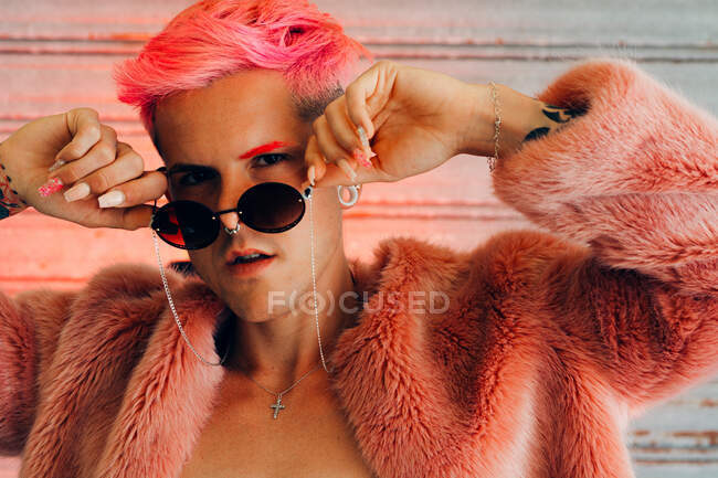 Junge Homosexuelle in stylischer Kleidung mit Tätowierung, rosa Haaren und Maniküre mit flauschiger Jacke und Sonnenbrille vor der Kamera — Stockfoto