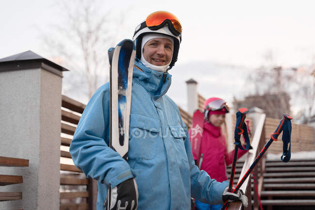 Щасливий батько і дочка носять теплий спортивний одяг і шоломи, що стоять з лижами на сніжному схилі пагорба і дивляться на камеру з задоволенням — стокове фото