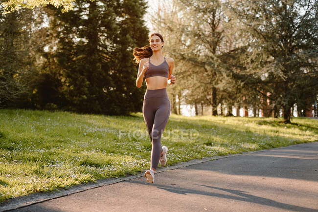 Atleta femenina alegre en ropa deportiva corriendo en carretera de asfalto mientras mira hacia otro lado durante el entrenamiento cardiovascular en la luz del sol - foto de stock