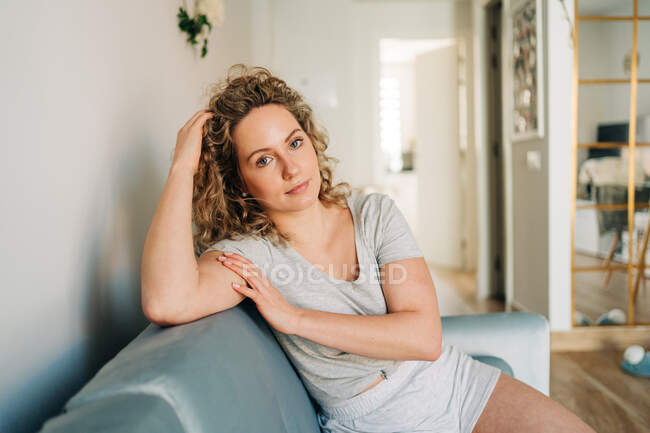 Positive attractive female in shorts sitting on cozy couch in living room and looking at camera — Foto stock