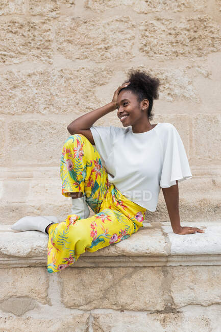 На виду усміхається афро-американська жінка в модному одязі сидячи навпроти стіни і озираючись у сонячне літо — стокове фото