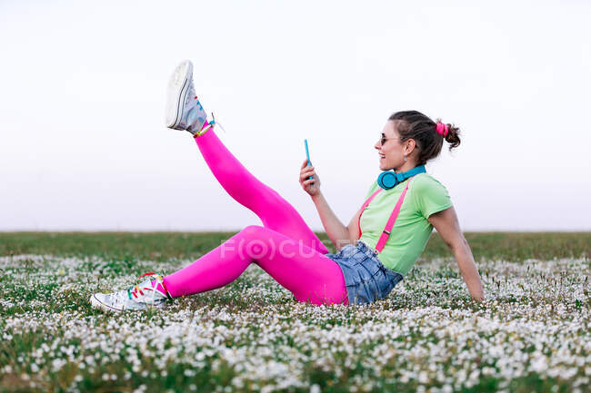 Side view full body joyful young female wearing bright outfit lying on lush grass with raised legs and browsing mobile phone in countryside — Stock Photo