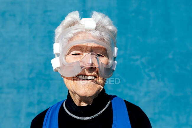 Cheerful mature female with gray hair wearing protective boxing headgear against blue wall and looking at camera with smile — Stock Photo