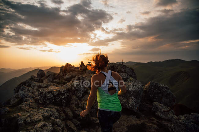Вид ззаду анонімної жінки в активному одязі, що стрибає на валуні високого хребта в долині гір під хмарним небом на заході сонця — стокове фото