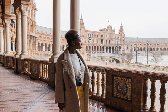 Giovane donna afroamericana in abbigliamento casual ammirare viste del grande palazzo con colonnato stucco ornamenti di lavoro e colonne durante la visita Siviglia Spagna — Foto stock