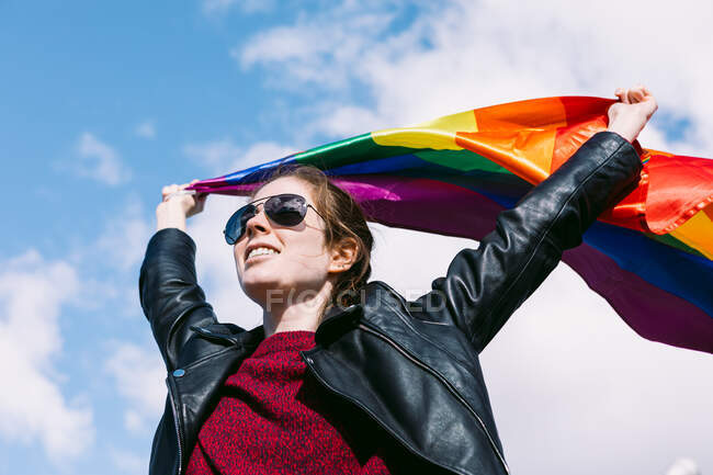 Знизу радісна лесбіянка стоїть на вулиці з ЛГБТ райдужним прапором, що пурхає вітром і озирається на хмарне небо. — стокове фото
