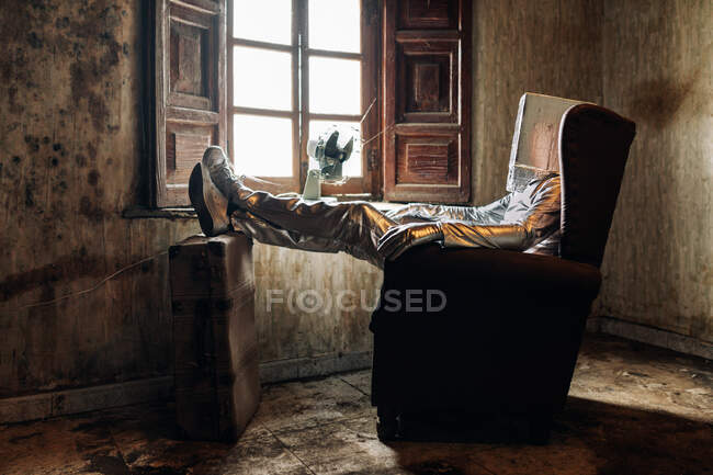 Vista lateral irreconocible persona con traje protector de plata con caja en la cabeza descansando en el sillón en la habitación en mal estado en casa abandonada - foto de stock