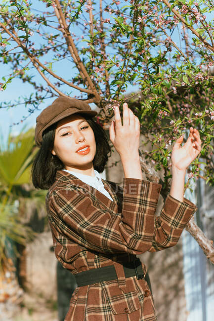 Attractive content brunette wearing trendy checkered coat and hat standing with hands raised in sunny spring garden and looking at camera - foto de stock