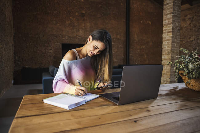 Content female remote employee with cellphone and netbook taking notes in notepad at table in house — Foto stock