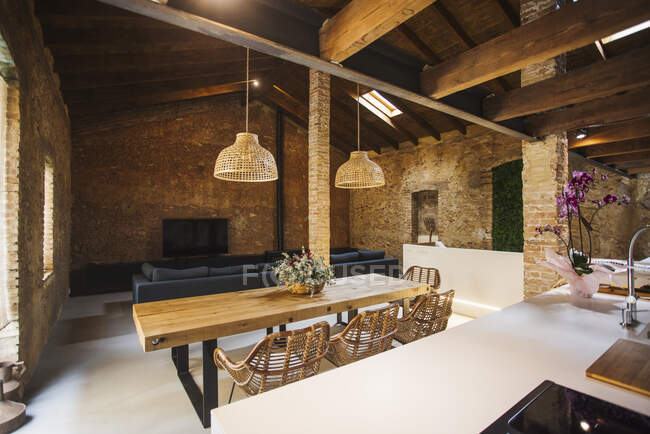 Kitchen and dining room interior with wooden table and wicker armchairs under lamps against brick walls in light house — Stock Photo