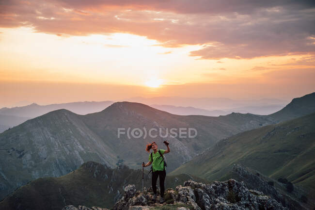 Trekker femenino con poste y teléfono de pie en el pico rocoso de la cresta de la montaña contra el cielo puesta del sol - foto de stock