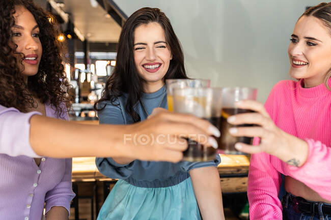 Joyful young multiethnic female best friends in stylish colorful outfits smiling and clinking glasses of refreshing juice and coke in modern bar — Stock Photo