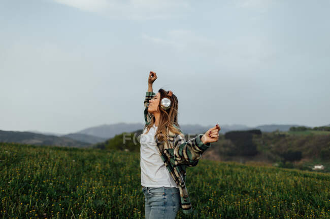 Side view of female listening to music from headphones while having fun dancing with eyes closed on summer meadow against mounts — Photo de stock