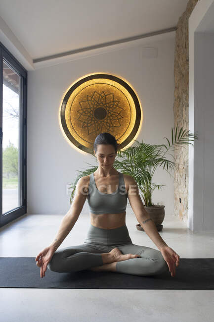 Сконцентрированная женщина в спортивной одежде сидит со скрещенными ногами и закрытыми глазами на коврик для йоги дома — стоковое фото