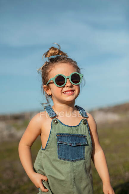 Zufriedenes kleines Mädchen in Overalls und Sonnenbrille steht auf der Wiese und genießt den Sommer an einem sonnigen Tag im Grünen — Stockfoto