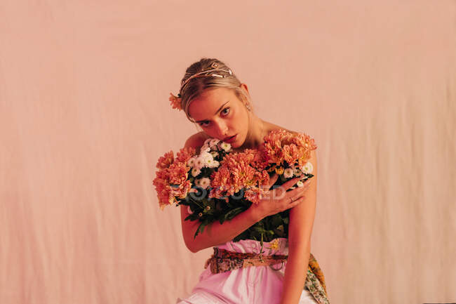 Encantadora joven romántica con hombros desnudos hembra con ramo de flores frescas en pie sobre fondo beige iluminado con luz de neón - foto de stock