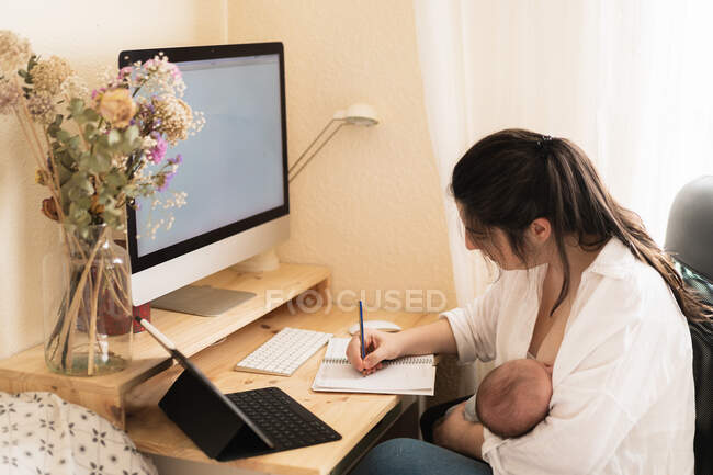 Adult mother feeding newborn with breast milk while taking notes in notebook at table in daytime — Stock Photo
