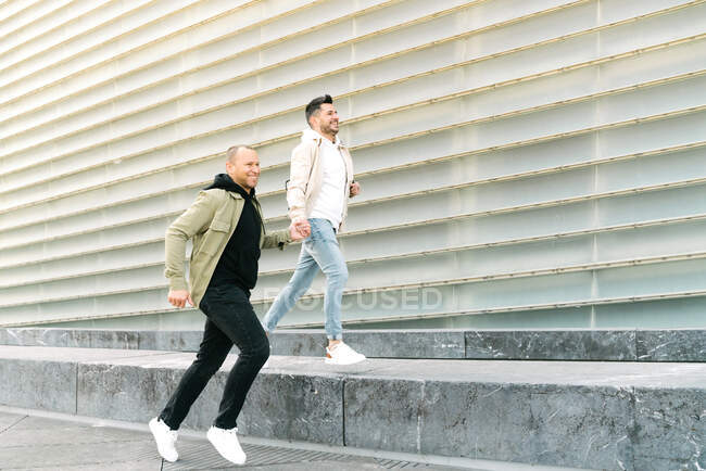 Side view of cheerful young multiracial gays in trendy outfits holding hands while running on paved street near modern building during romantic date in city — Foto stock