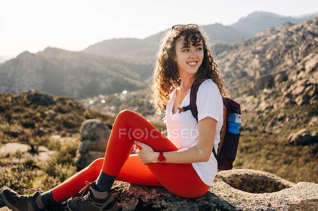 Vue latérale d'une jeune voyageuse joyeuse aux cheveux noirs bouclés en tenue décontractée souriant et détournant les yeux tout en recréant sur pierre lors d'un voyage de randonnée en montagne — Photo de stock