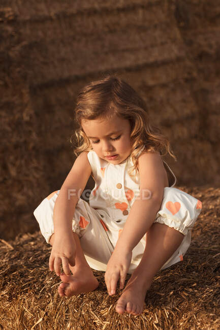Fröhlich liebenswertes Kind in Overalls, das mit Heu spielt und auf Strohballen in der Landschaft sitzt — Stockfoto