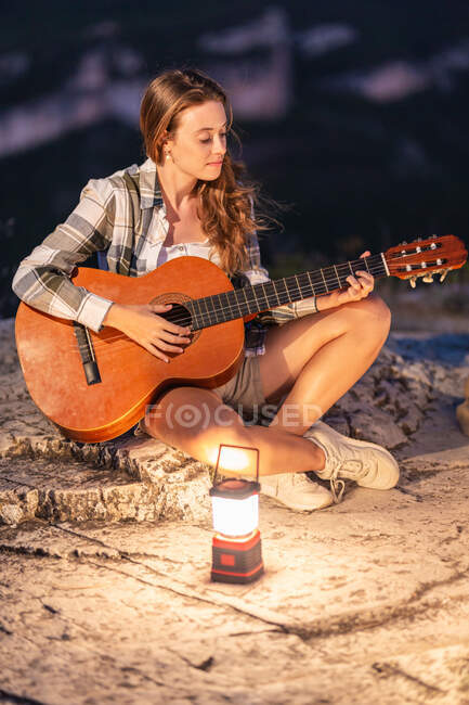 Высокий угол женщины-путешественницы, сидящей на скале с светящимся фонарем и играющей на акустической гитаре, наслаждаясь вечером в высокогорье — стоковое фото