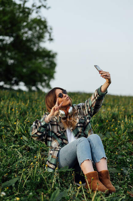 Young female in sunglasses with headphones showing peace gesture while taking self portrait on cellphone and sitting on meadow — Fotografia de Stock