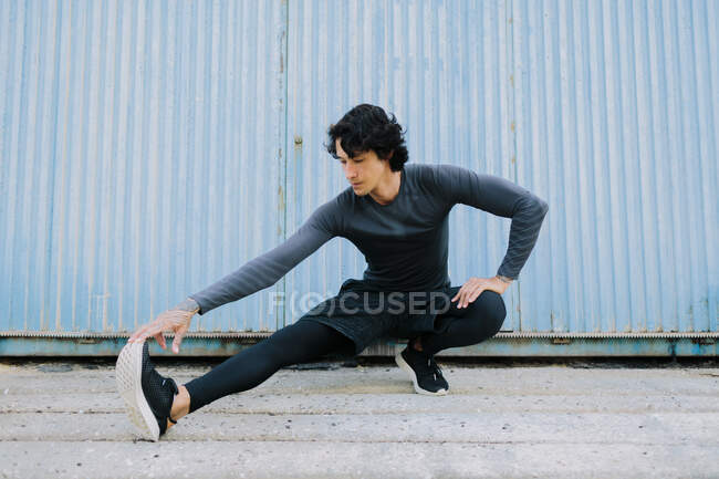Full body young ethnic male in sportswear sitting on haunches and warming up legs on street pavement — Stock Photo