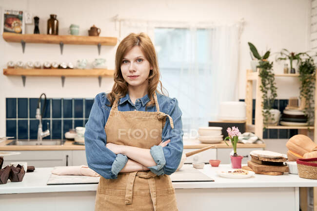 Молодая самоуверенная женщина со сложенными руками в фартуке смотрит в камеру дома — стоковое фото