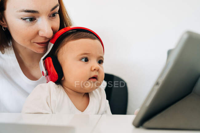 Positiva joven madre y lindo bebé atento en los auriculares viendo dibujos animados en la tableta mientras están sentados en el escritorio juntos - foto de stock